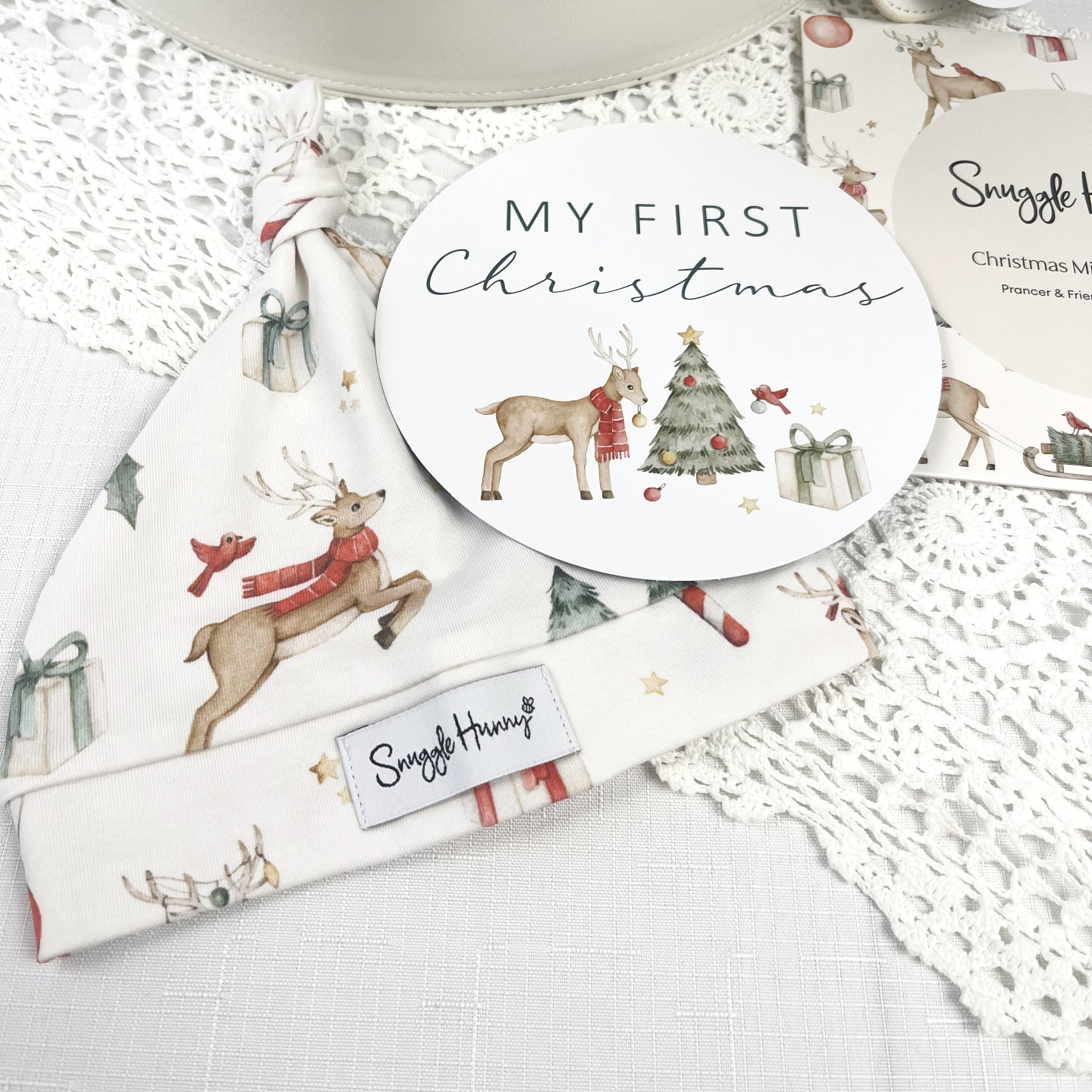 Baby beanie and milestone card with Christmas-themed design on a white lace background