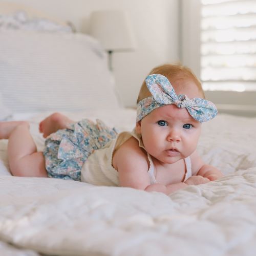 baby dressed in liberty blue print frilly romper and headband laying on bed with white duvet