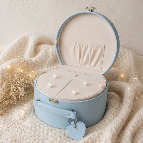 A light bluekeepsake box with its lid open, showing a cream-colored interior organiser and a small heart-shaped tag. The box is styled on a textured cream knit blanket with warm fairy lights and dried flowers.