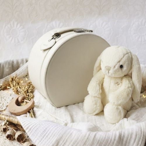 A cream-colored keepsake box with a handle and clasp, styled next to a plush cream bunny toy on a white textured blanket with dried floral accents.