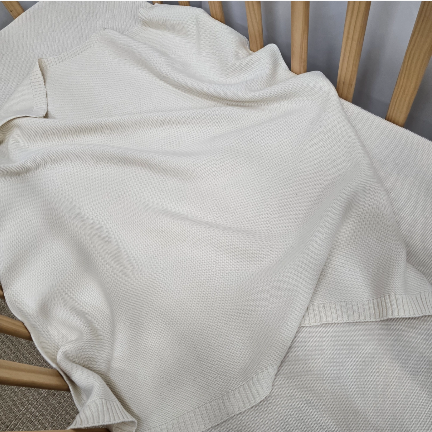 A cream-colored knit blanket draped over a light wood slatted chair, showing its ribbed cuffs and hem.