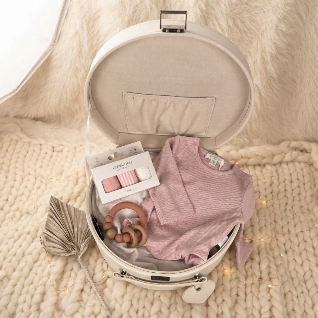 An open cream-colored keepsake box styled as a nursery gift set, containing a dusty pink baby suit, a wooden teething ring, and a pack of baby socks, set on a chunky knit blanket.