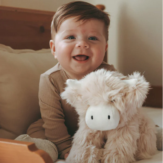 Child sitting on a bed with a fluffy highland cow toy, smiling.