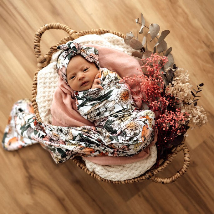 Newborn baby wrapped in a floral blanket with dried flowers in a woven basket on a wooden floor.