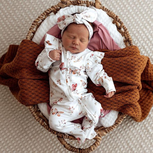 Newborn baby in a butterfly print outfit and headband, wrapped in brown blanket, lying in a woven basket.