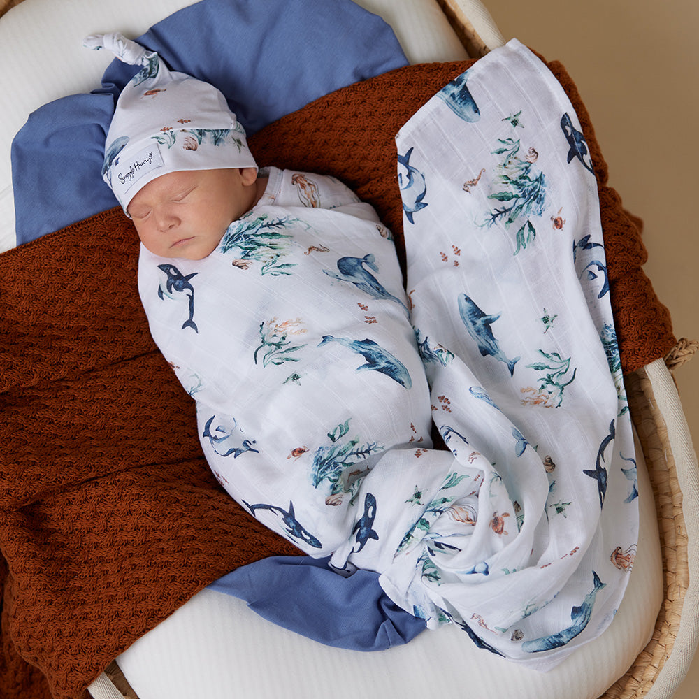 Newborn baby wrapped in a muslin wrap with whale design, wearing a matching hat, in a wooden crib.