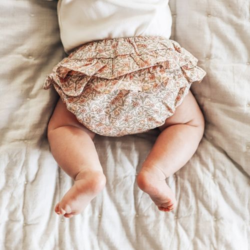 A baby lying on a quilted cream blanket, wearing a ruffled bloomers-style diaper cover with a delicate floral pattern in muted pink, rust, and beige tones.