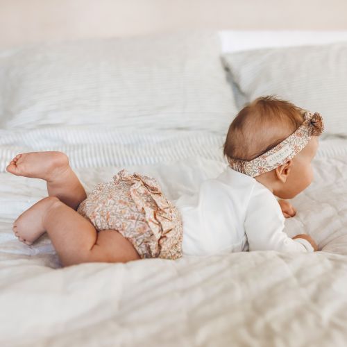A baby lying on a cream-colored quilted bed, wearing a white long-sleeve bodysuit, matching patterned ruffled diaper cover, and a floral patterned headband.