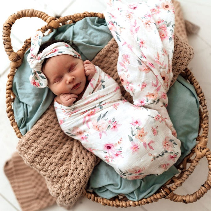 Newborn baby wrapped in floral blanket and headband, lying in a woven basket with soft textures.