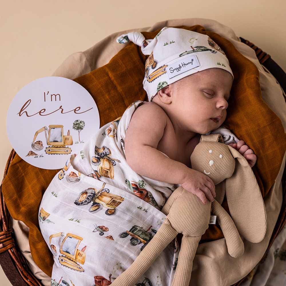 Newborn baby in a basket with a toy, wearing a hat and swaddle with vehicle prints.
