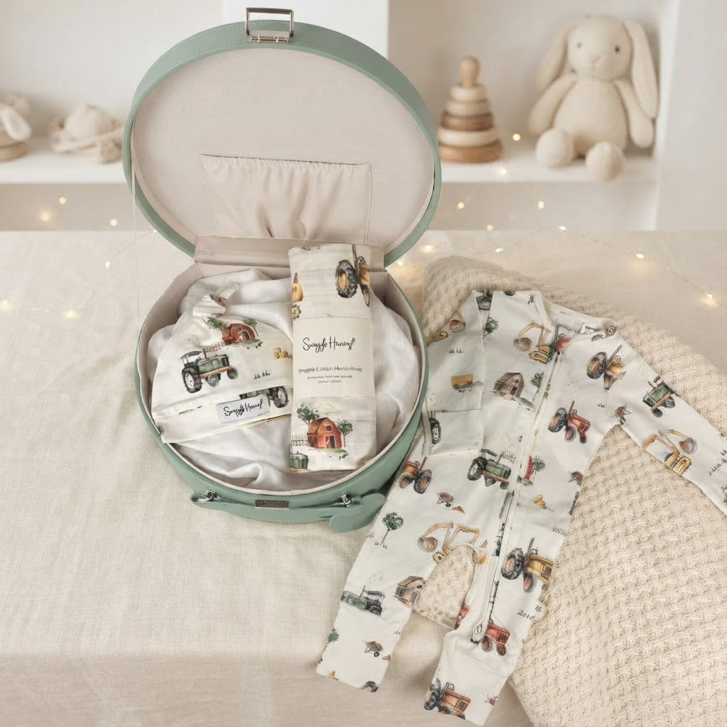 open keepsake box with digger print baby onesie, beanie and wrap laying on a bed in a baby nursery with toys on a shelf behind
