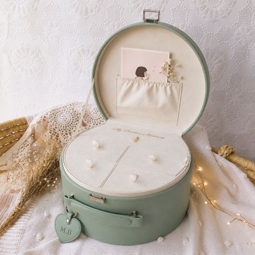 A sage green keepsake box shown open, with a cream-colored interior, a card pocket, and a hanging heart-shaped tag. The box is styled on a white textured fabric with dried flowers and warm string lights in the background.
