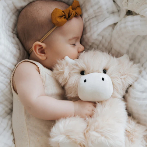 Sleeping baby hugging a cream highland cow plush toy on neutral background