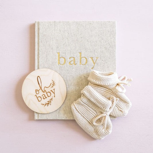 Knitted baby booties and a wooden disc with 'oh baby' text on a light pink background