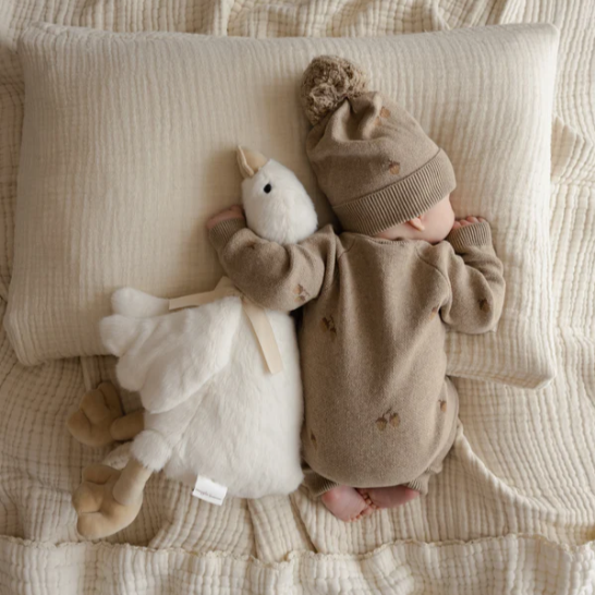 Baby in a brown outfit with a matching hat lying on a textured surface next to a Snuggle Bunny plush toy duck.