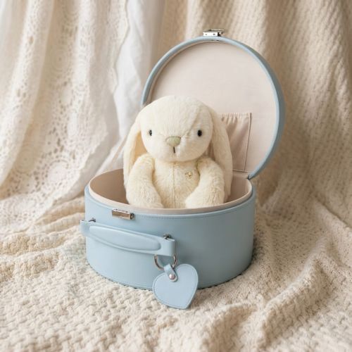 A cream-colored plush bunny toy sits inside an open light blue keepsake box with a handle and heart-shaped tag, placed on a textured cream knit blanket.