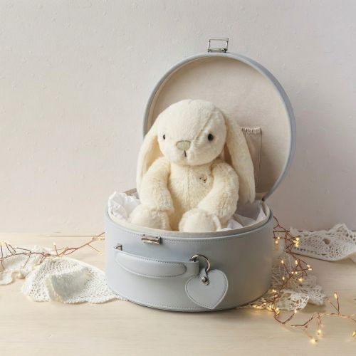 A cream-colored plush bunny stuffed animal sitting inside an open grey keepsake box, styled on a light wood surface with lace fabric and warm string lights against a soft beige wall.