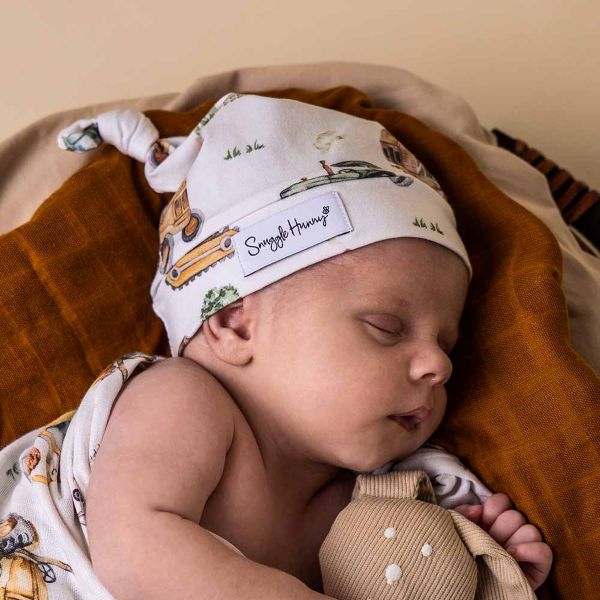 Newborn baby wearing a 'Snuggle Hunny' hat with a pattern of cars and trees, lying on a brown blanket.