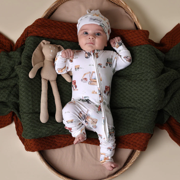 Baby in a digger patterned onesie and hat laying in a basket with a soft toy, surrounded by green and brown blankets.
