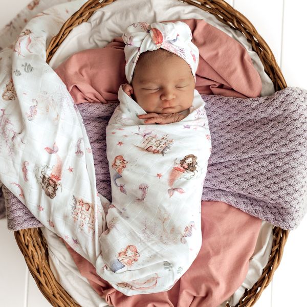 Newborn baby wrapped in a mermaid print wrap with a headband, lying in a wicker basket.