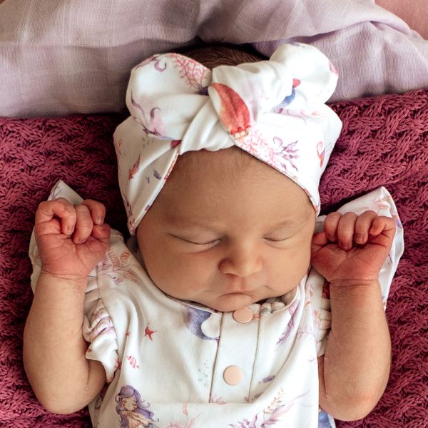 Newborn baby wearing a mermaid print headband and outfit, sleeping on a pink blanket.
