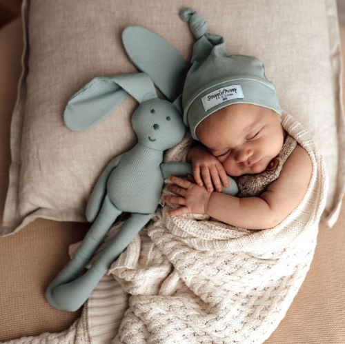 sleeping baby wrapped in a cream blanket wearing a sage knotted beanie next to a sage snuggle bunny toy