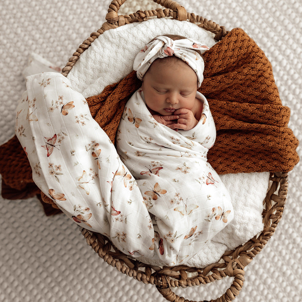 Newborn baby wrapped in a floral blanket with a brown wrap, lying in a wicker basket on a textured surface.