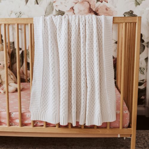 Wooden crib with a white knitted blanket draped over it, floral wallpaper in the background